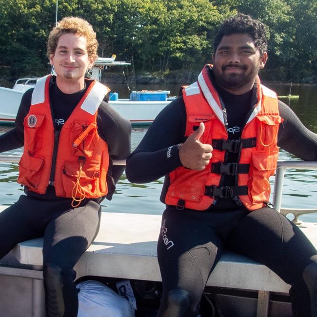 Two students in life vests smile as they sit on a bench of a U N E boat